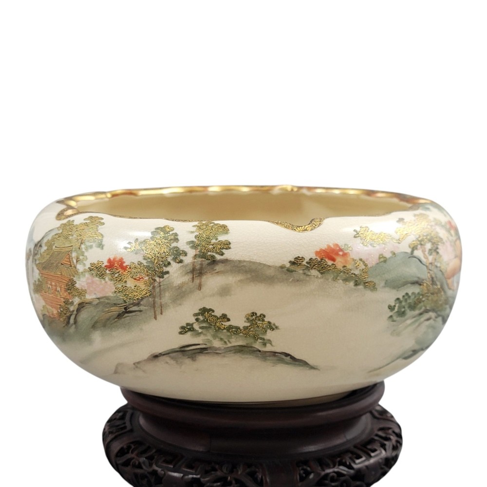 A Japanese Satsuma earthenware bowl with hand-painted figures under trees and gold accents, resting on a dark carved wooden stand. View XI