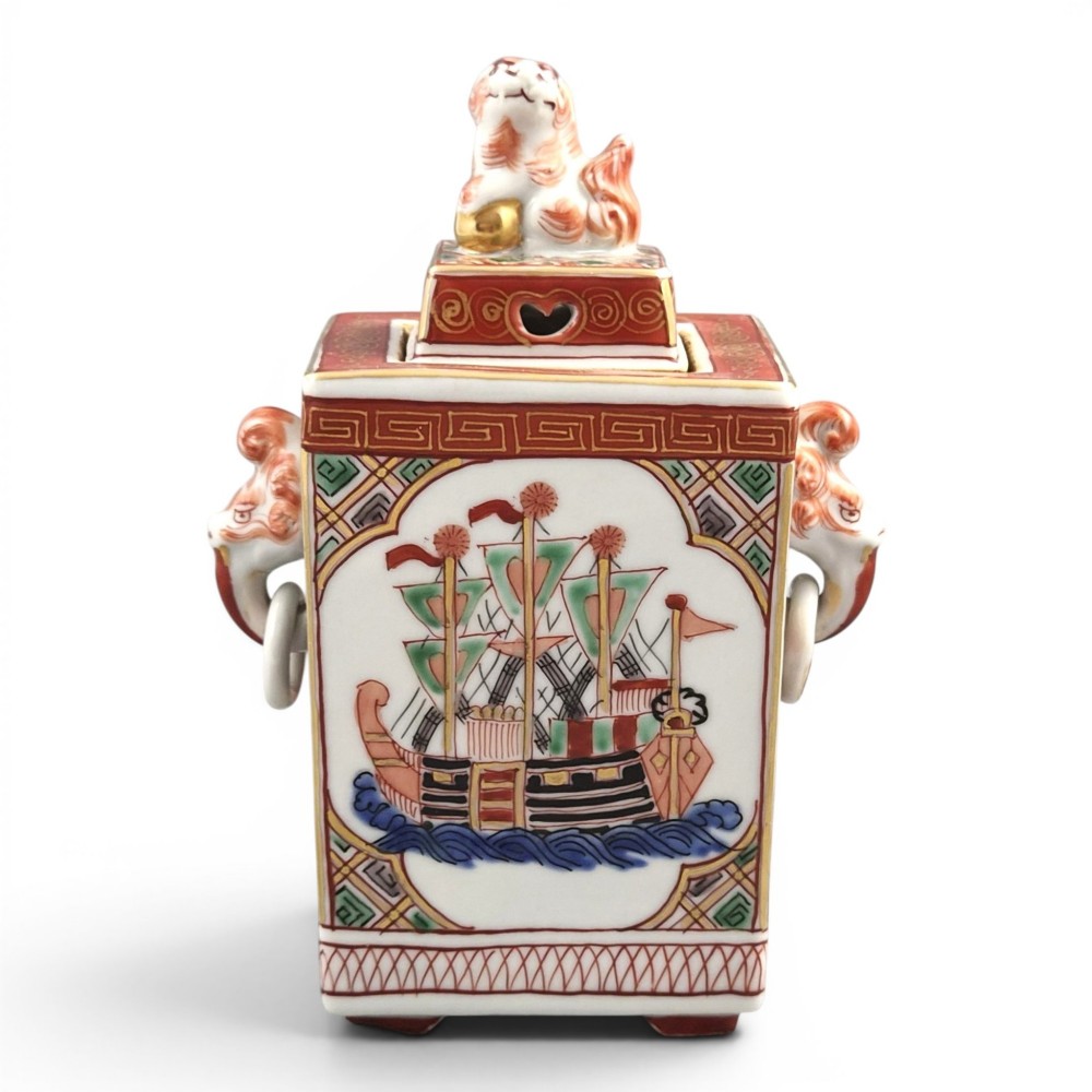A rectangular Japanese porcelain incense burner featuring hand-painted depictions of a European ship, decorative red enamel borders, animal head handles, and a shishi lion finial on the lid.