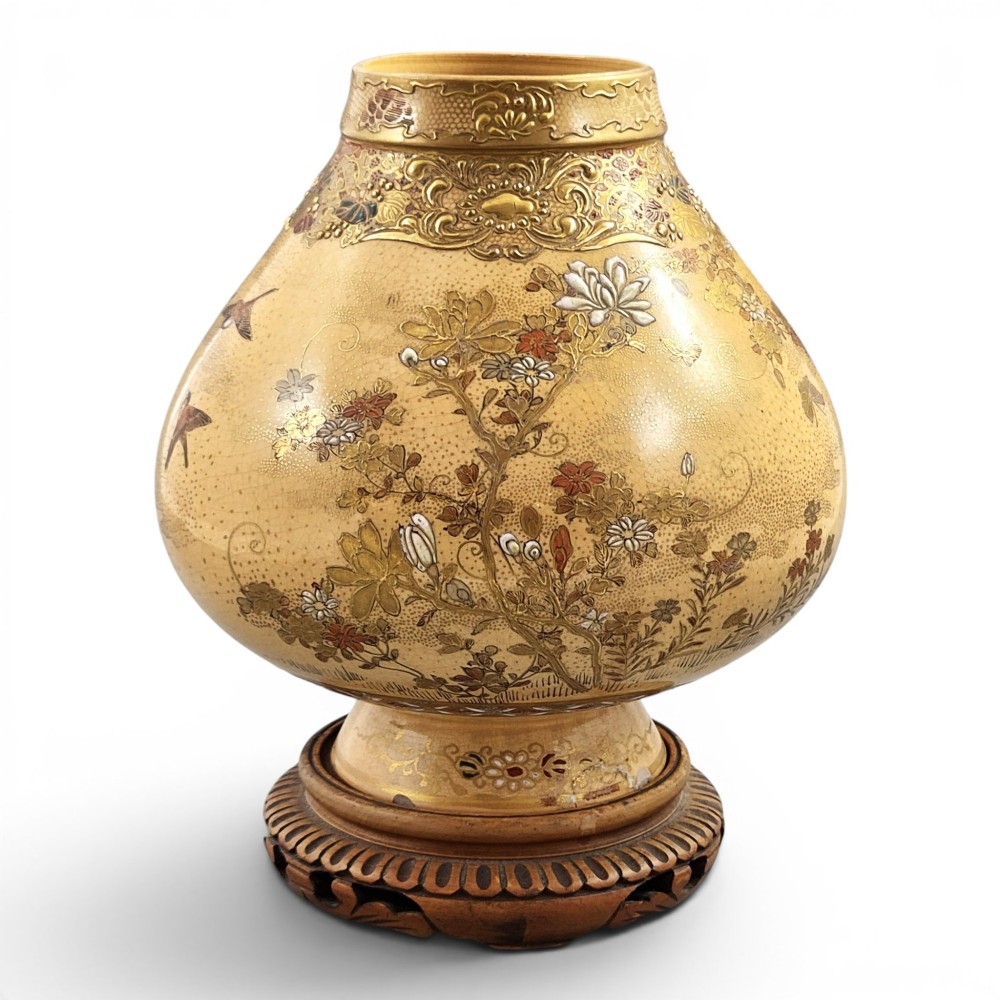 A bulbous Japanese Satsuma earthenware vase featuring a cream crackle glaze, intricate floral gilding, birds, and a carved dark wooden display stand.