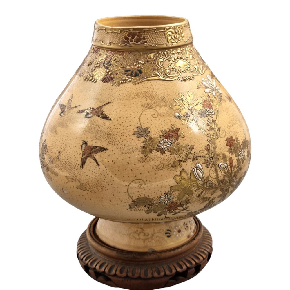 A bulbous Japanese Satsuma earthenware vase featuring a cream crackle glaze, intricate floral gilding, birds, and a carved dark wooden display stand. View X