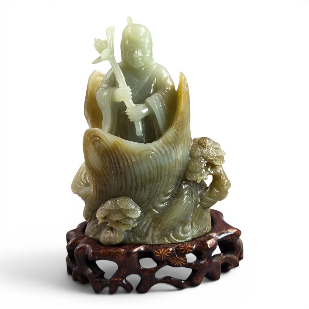 A carved pale green Chinese jadeite figure holding a lotus stem, resting on a fitted dark carved wooden stand.