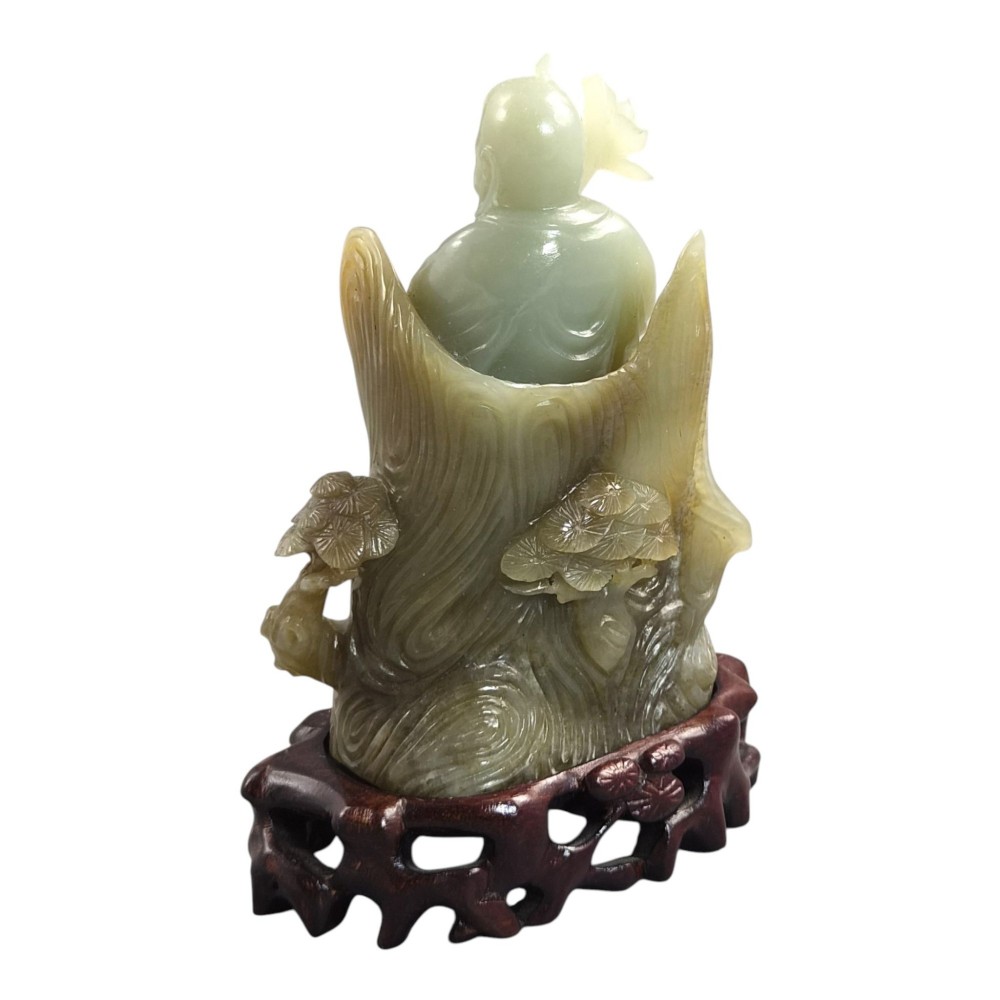 A carved pale green Chinese jadeite figure holding a lotus stem, resting on a fitted dark carved wooden stand. View IV