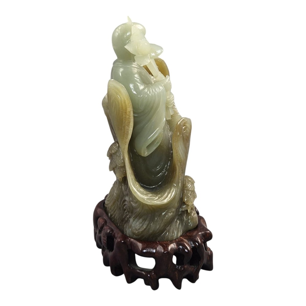 A carved pale green Chinese jadeite figure holding a lotus stem, resting on a fitted dark carved wooden stand. View VI