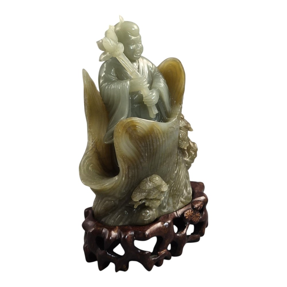 A carved pale green Chinese jadeite figure holding a lotus stem, resting on a fitted dark carved wooden stand. View VII