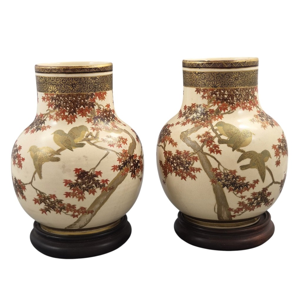 A pair of bulbous cream-coloured Japanese Satsuma earthenware vases decorated with gilt birds and autumnal foliage on dark carved wood stands.