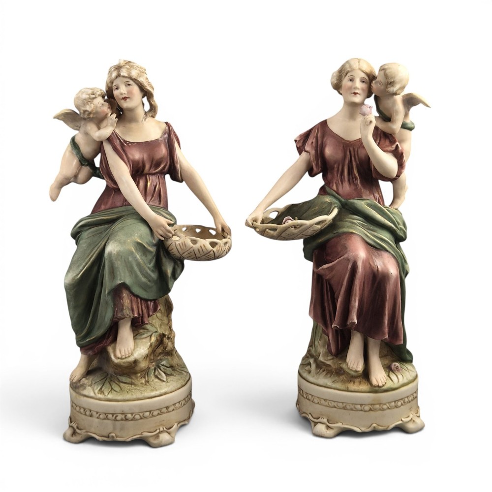 A pair of Royal Dux Art Nouveau porcelain figurines depicting classical maidens with winged cherubs and openwork baskets.