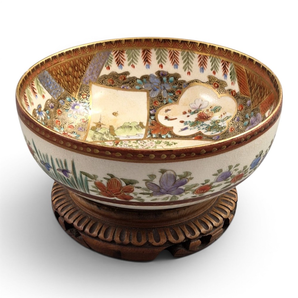 An antique Meiji period Japanese Satsuma earthenware bowl featuring hand-painted floral panels and gold detailing, elevated on a carved dark wooden base.