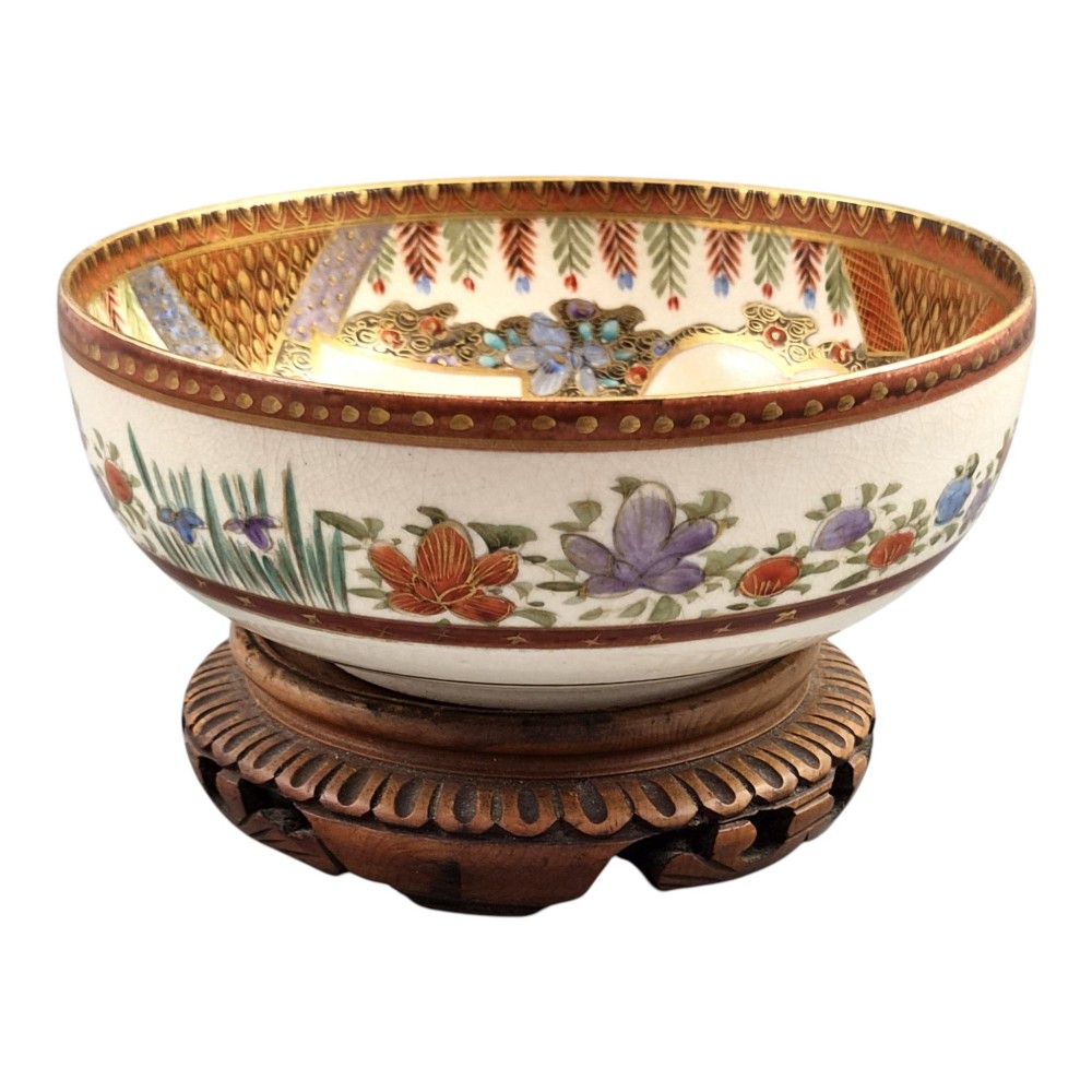 An antique Meiji period Japanese Satsuma earthenware bowl featuring hand-painted floral panels and gold detailing, elevated on a carved dark wooden base. View III