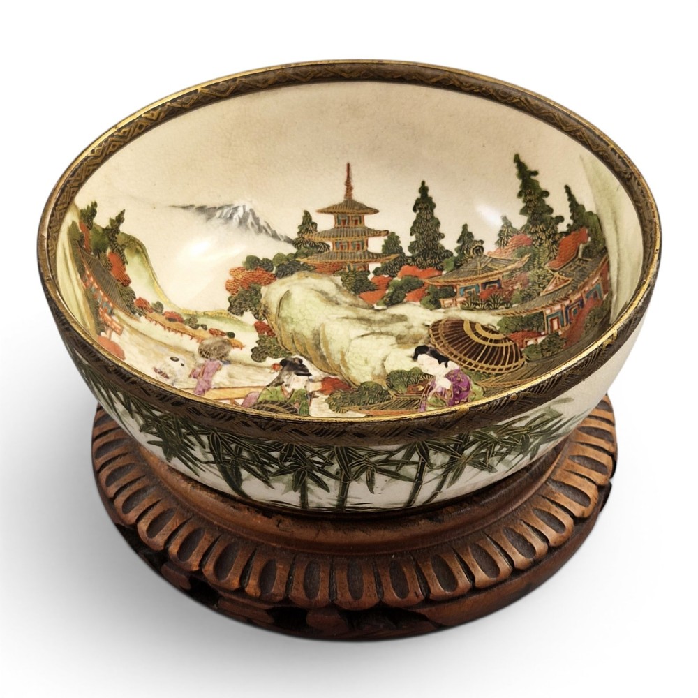 An antique cream earthenware bowl resting on a carved wooden base, decorated inside with hand-painted Japanese landscapes and figures, and finished with a bamboo pattern around the exterior.