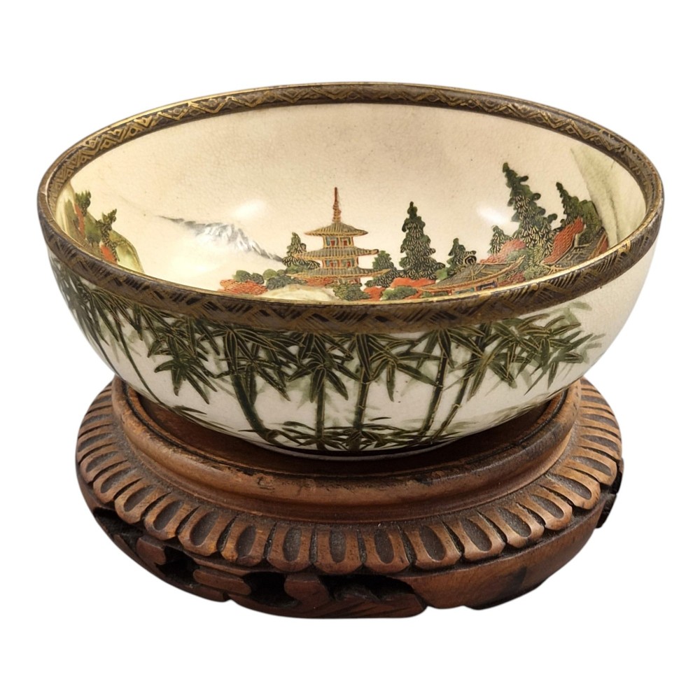 An antique cream earthenware bowl resting on a carved wooden base, decorated inside with hand-painted Japanese landscapes and figures, and finished with a bamboo pattern around the exterior. View X