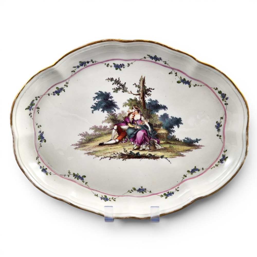 An oval 18th-century French porcelain tray featuring a hand-painted pastoral scene bordered by pink ribbon decoration and a scalloped gilt edge.