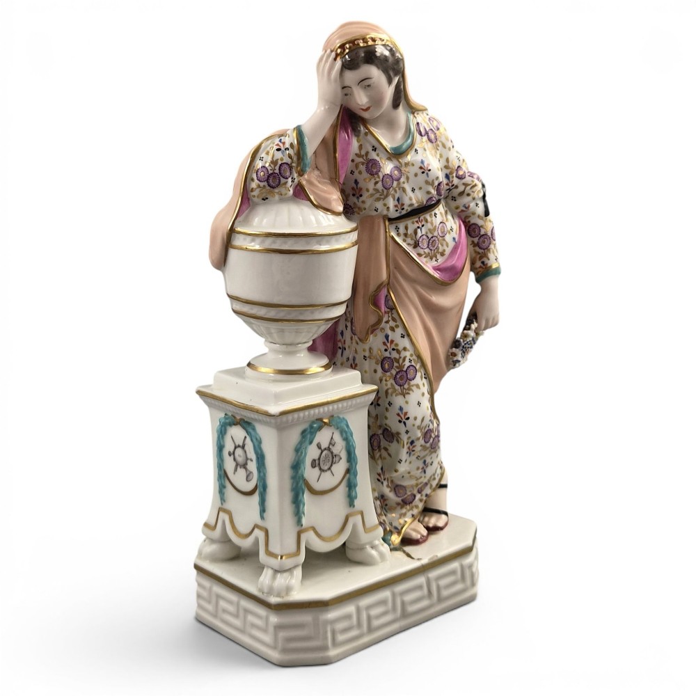 A Neoclassical porcelain figure depicting a draped female leaning against a gilded urn on a pedestal.