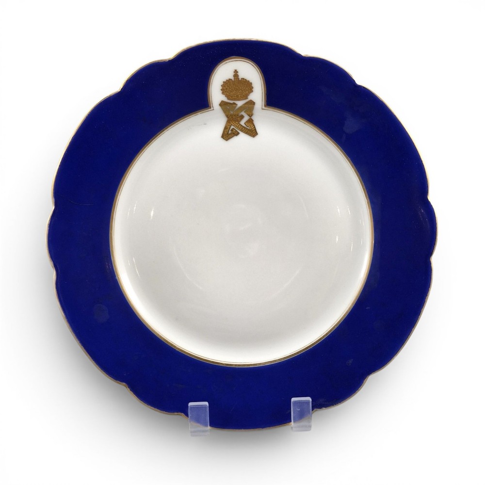 A white round porcelain plate featuring a thick cobalt blue scalloped border and a gilded Imperial monogram topped with a crown.