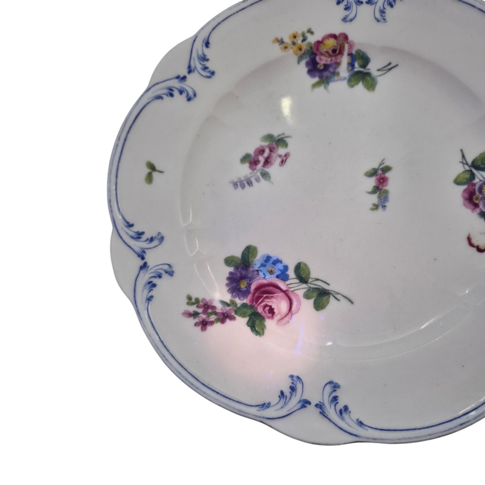 An 18th-century circular white soft-paste porcelain plate decorated with hand-painted floral sprays and a scalloped rim with blue feathered scroll details. View X
