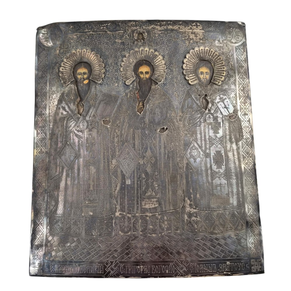 A rectangular antique Orthodox silver icon featuring repousse depictions of the Three Holy Hierarchs with detailed halos and Cyrillic script along the bottom edge.