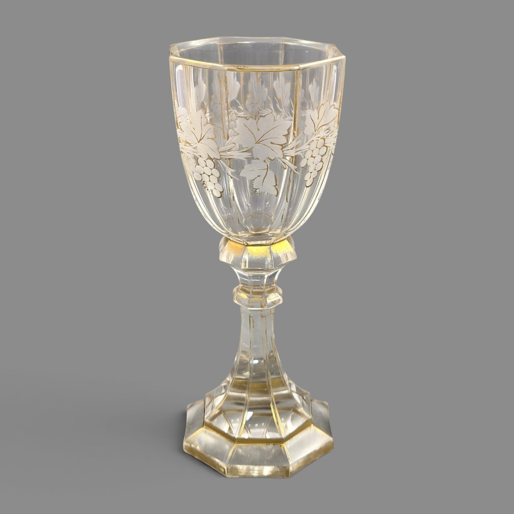 A clear Bohemian crystal romer goblet featuring wheel-engraved grapevine motifs, a faceted stem, and gilt highlight accents on a hexagonal foot. View VI