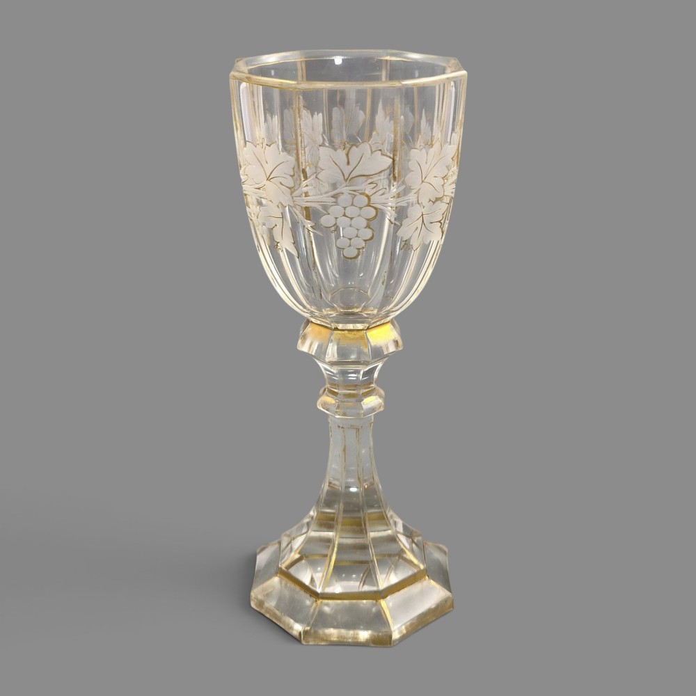 A clear Bohemian crystal romer goblet featuring wheel-engraved grapevine motifs, a faceted stem, and gilt highlight accents on a hexagonal foot. View VII