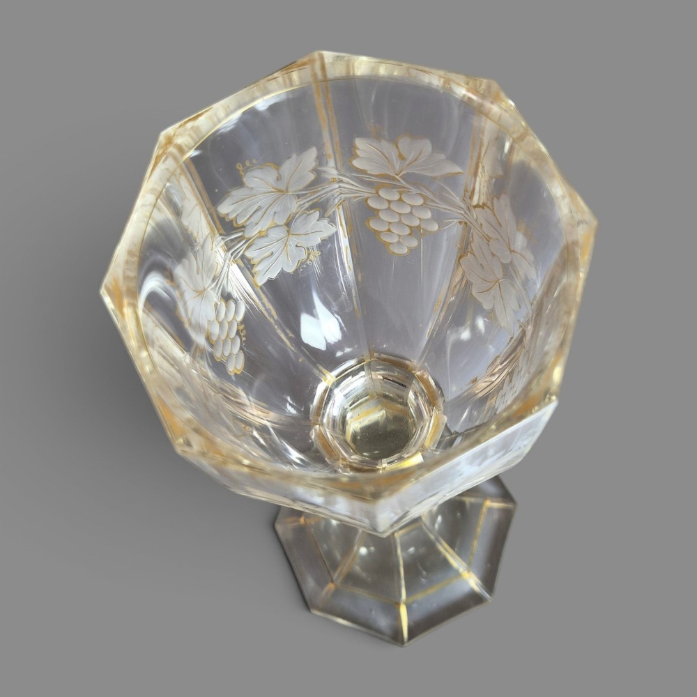 A clear Bohemian crystal romer goblet featuring wheel-engraved grapevine motifs, a faceted stem, and gilt highlight accents on a hexagonal foot. View IX