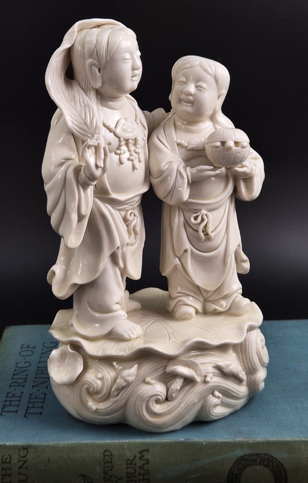 A cream white glazed porcelain figure group featuring two standing figures positioned on a lotus leaf and wave carved base.