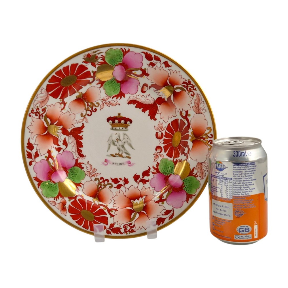 A circular white porcelain plate featuring a gilded rim, a red and pink floral border, and a central armorial crest of a bird beneath a coronet with a banner reading STRIKE. View VI