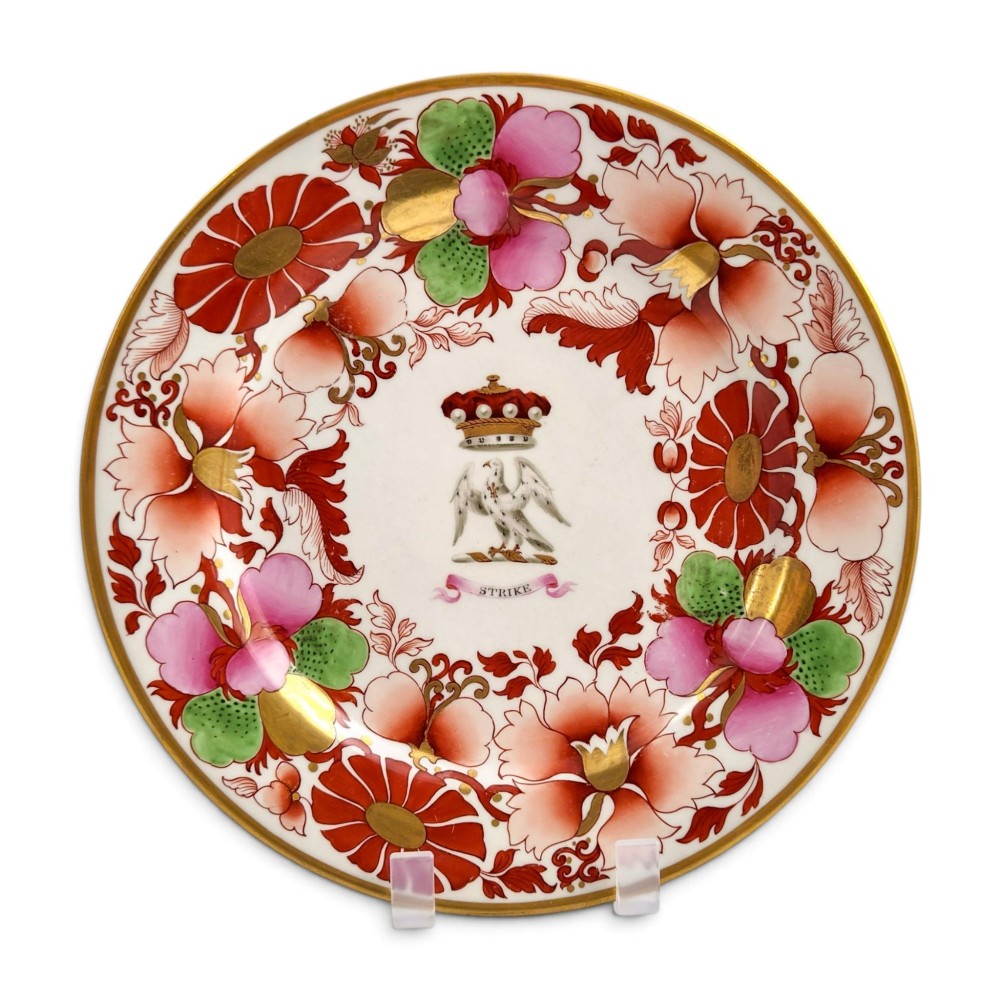 A circular white porcelain plate featuring a gilded rim, a red and pink floral border, and a central armorial crest of a bird beneath a coronet with a banner reading STRIKE.