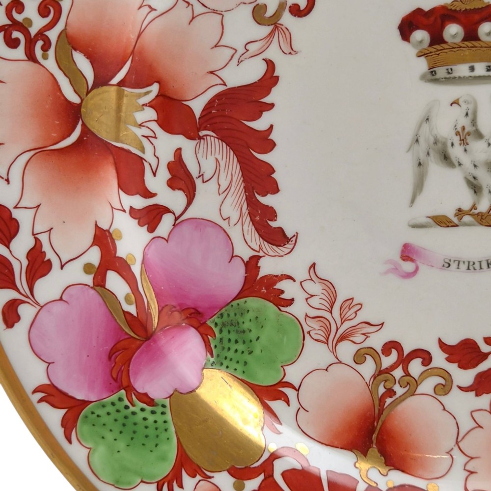 A circular white porcelain plate featuring a gilded rim, a red and pink floral border, and a central armorial crest of a bird beneath a coronet with a banner reading STRIKE. View IX