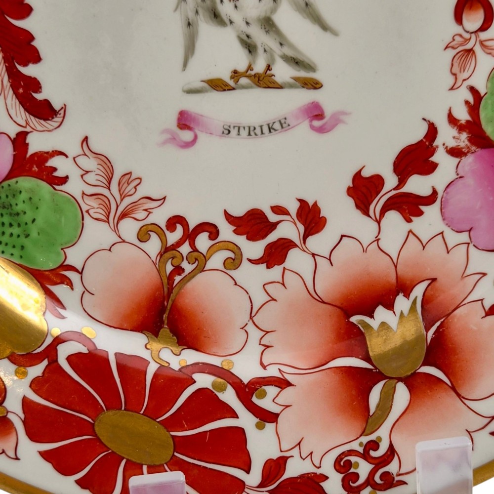 A circular white porcelain plate featuring a gilded rim, a red and pink floral border, and a central armorial crest of a bird beneath a coronet with a banner reading STRIKE. View X