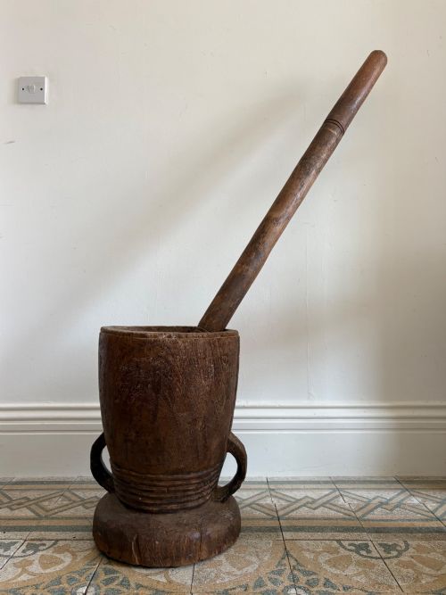 fig 1. A hand-carved African hardwood grain mortar with side loop handles and concentric carved grooves, next to a tall wooden pestle pole.
