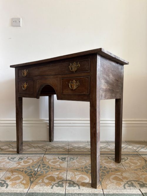 fig 2. An early eighteenth-century oak lowboy side table featuring one wide upper drawer, two small side drawers, an arched apron, brass handles, and straight square legs.