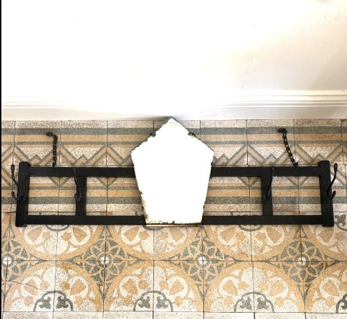 fig 2. A tall black wrought iron wall-mounted coat rack featuring integrated hooks, hanging chains, and an asymmetrical pentagonal mirror.
