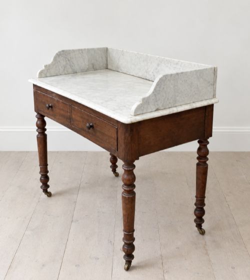 fig 2. A 19th-century mahogany washstand featuring a white Carrara marble top with splashback, two frieze drawers with wooden knobs, and turned legs with brass castors.