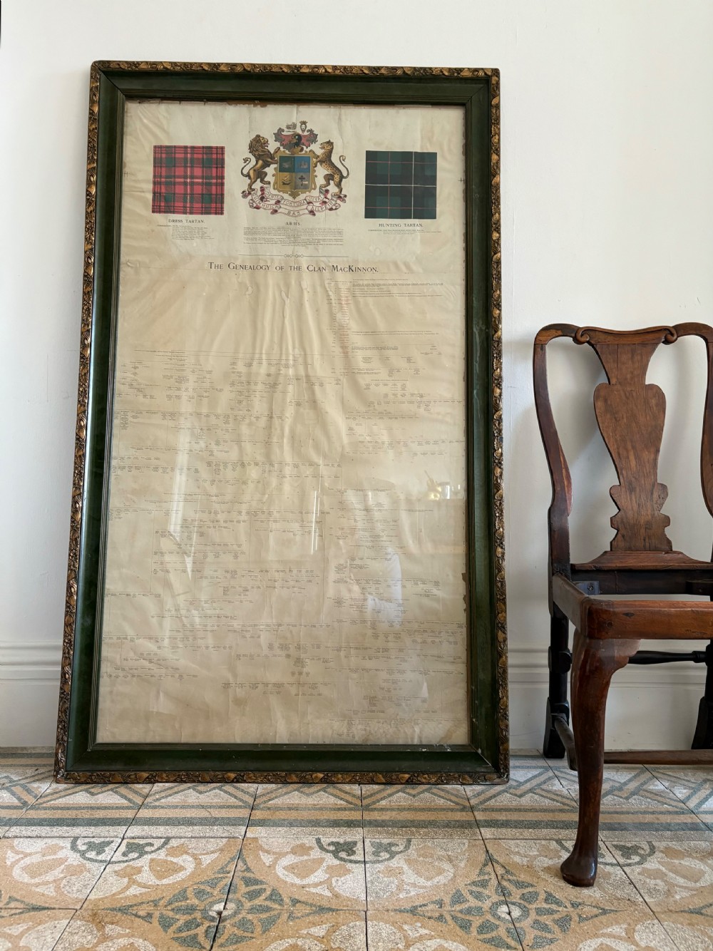 A large, vertically orientated framed paper genealogy chart for Clan Mackinnon featuring two tartan swatches and a central heraldic coat of arms at the top within a dark green and gilt wood frame. View IV
