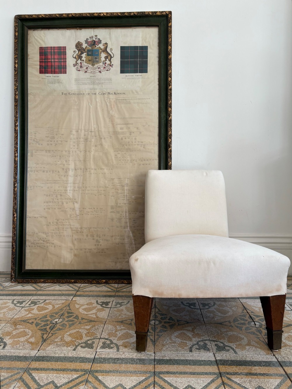 A large, vertically orientated framed paper genealogy chart for Clan Mackinnon featuring two tartan swatches and a central heraldic coat of arms at the top within a dark green and gilt wood frame. View XIII