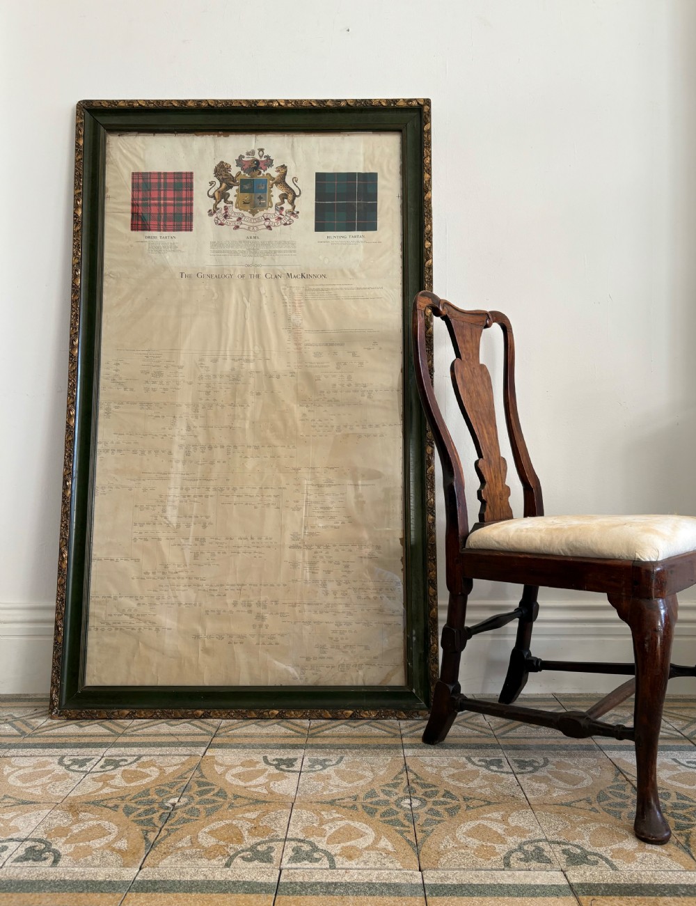 A large, vertically orientated framed paper genealogy chart for Clan Mackinnon featuring two tartan swatches and a central heraldic coat of arms at the top within a dark green and gilt wood frame. View XIV