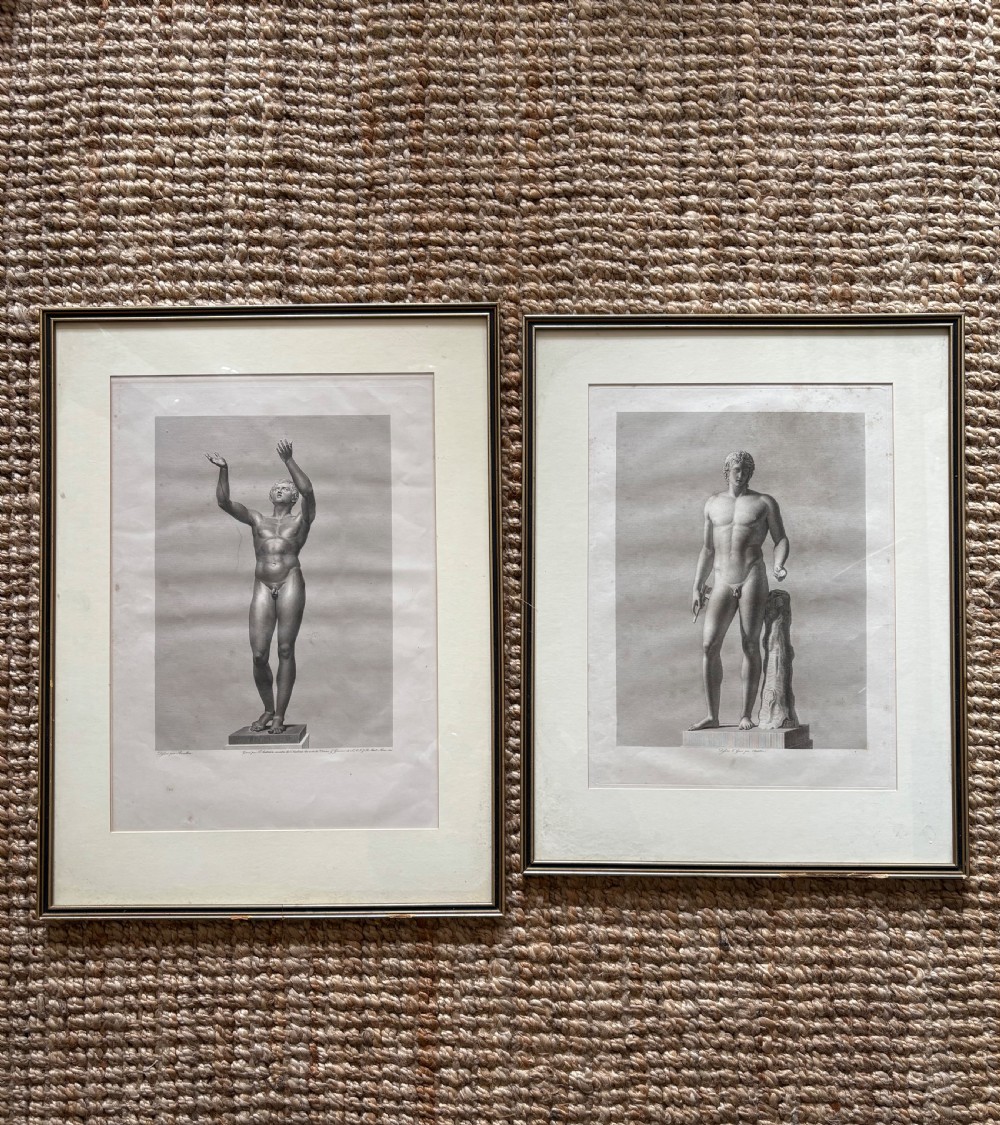 A pair of framed monochrome 19th-century classical engravings depicting ancient male sculptures mounted in cream mats.