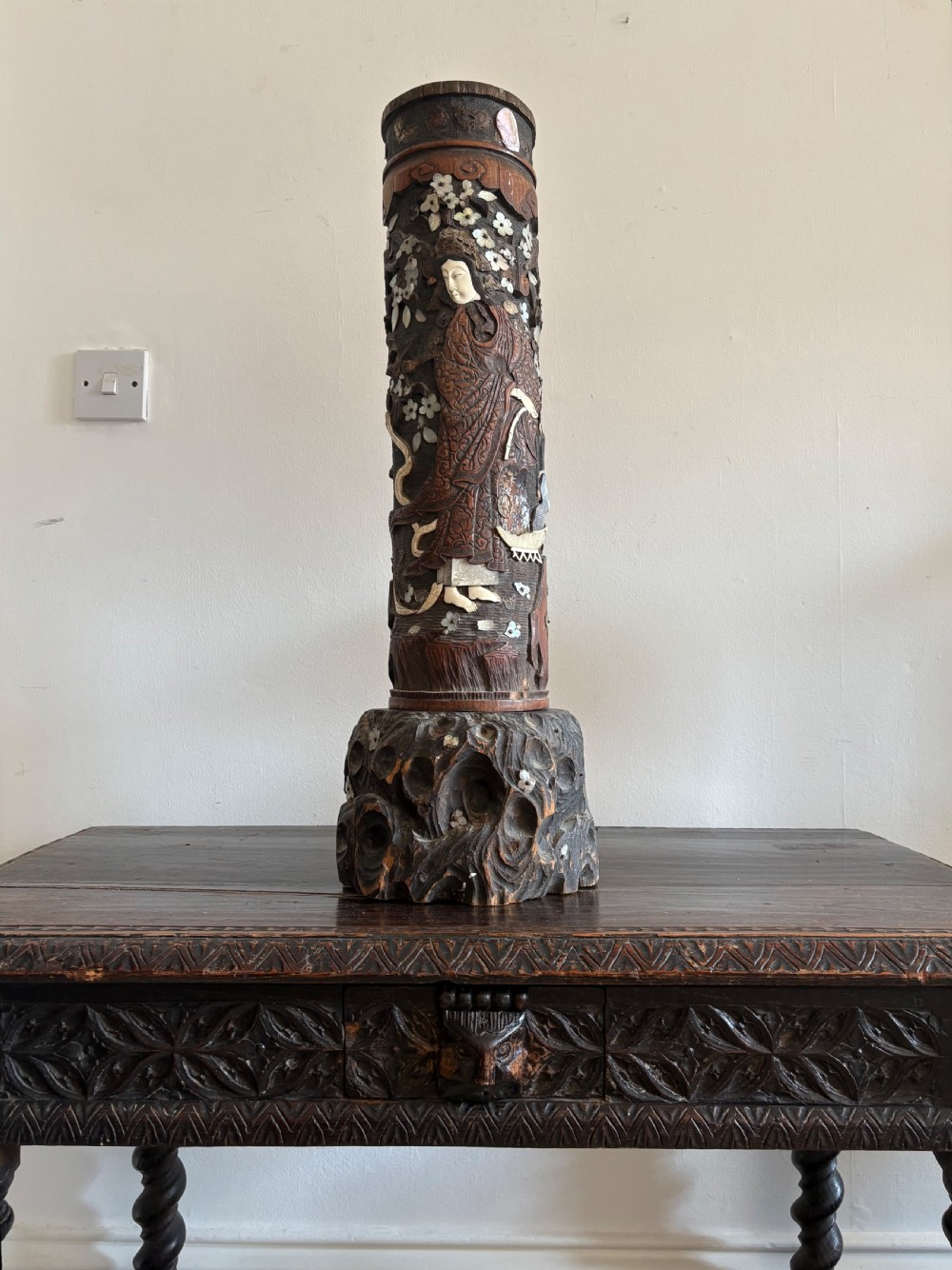 A tall 19th-century Japanese Meiji period carved wood brush pot featuring high-relief figures inlaid with mother-of-pearl and hardstone resting on an organic carved wood base.