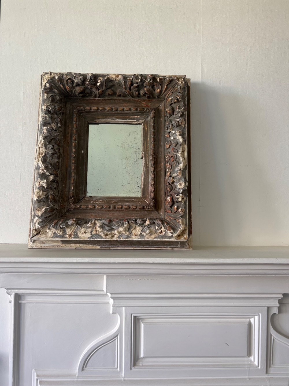 A small rectangular dark wood frame with heavily textured gesso carving enclosing a foxed mirror plate.