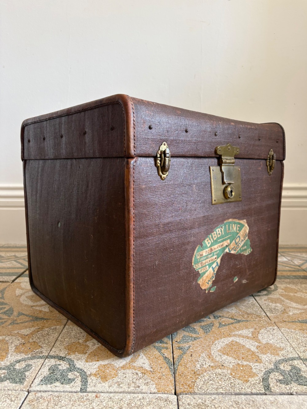 An Edwardian brown canvas and leather hat box trunk featuring brass latches, stitched leather trim, and an ocean liner travel label on the front.