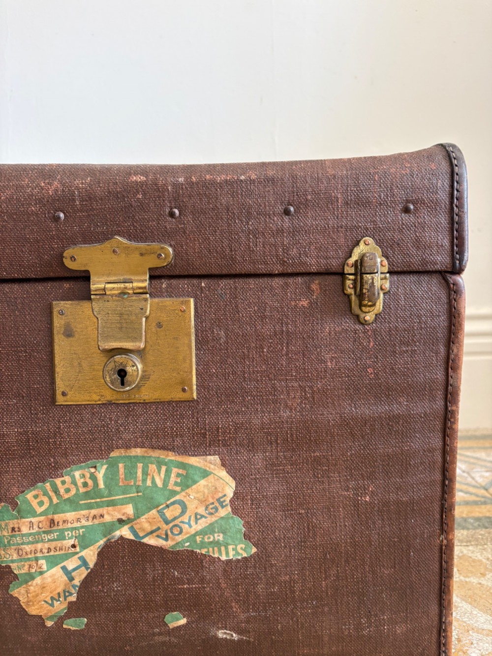 An Edwardian brown canvas and leather hat box trunk featuring brass latches, stitched leather trim, and an ocean liner travel label on the front. View X
