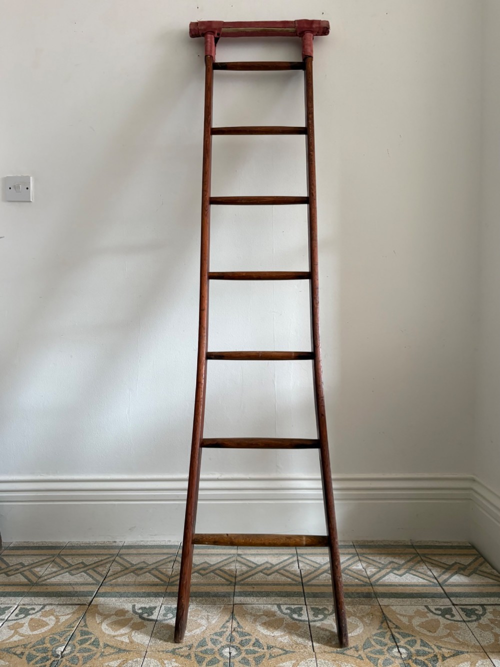 A tall early 20th-century pitch pine and elm library ladder with turned wooden rungs and a padded upper rail leaning against a white wall.