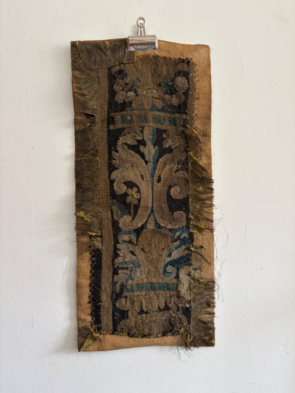 A vertical fragment of an antique hand-woven verdure tapestry featuring acanthus leaf motifs, mounted onto a natural linen backing.