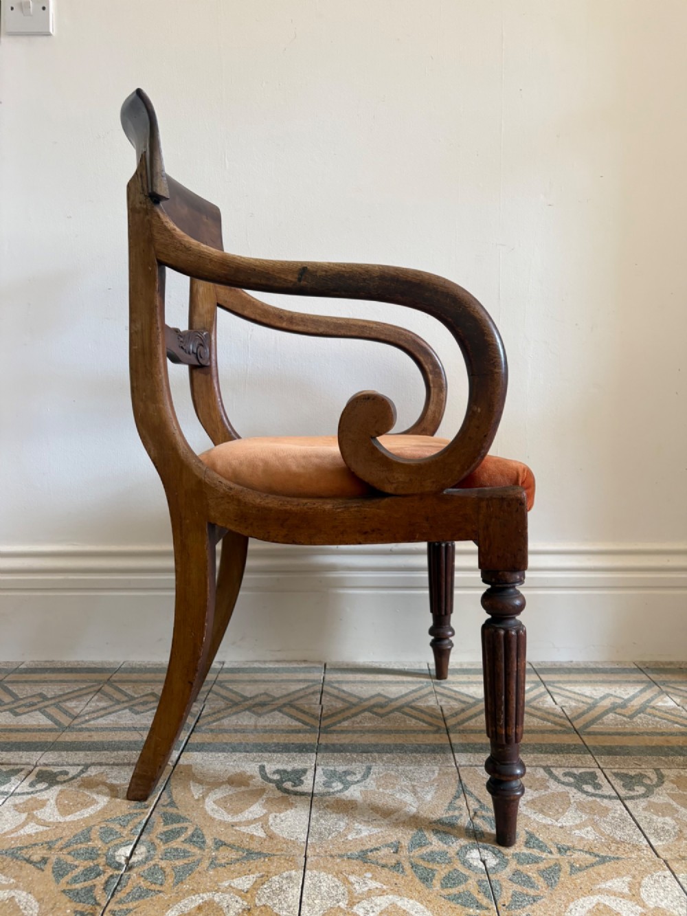 Profile view of a Late Regency mahogany open armchair with scrolled armrests, reeded front legs, and a rust orange velvet drop-in seat.