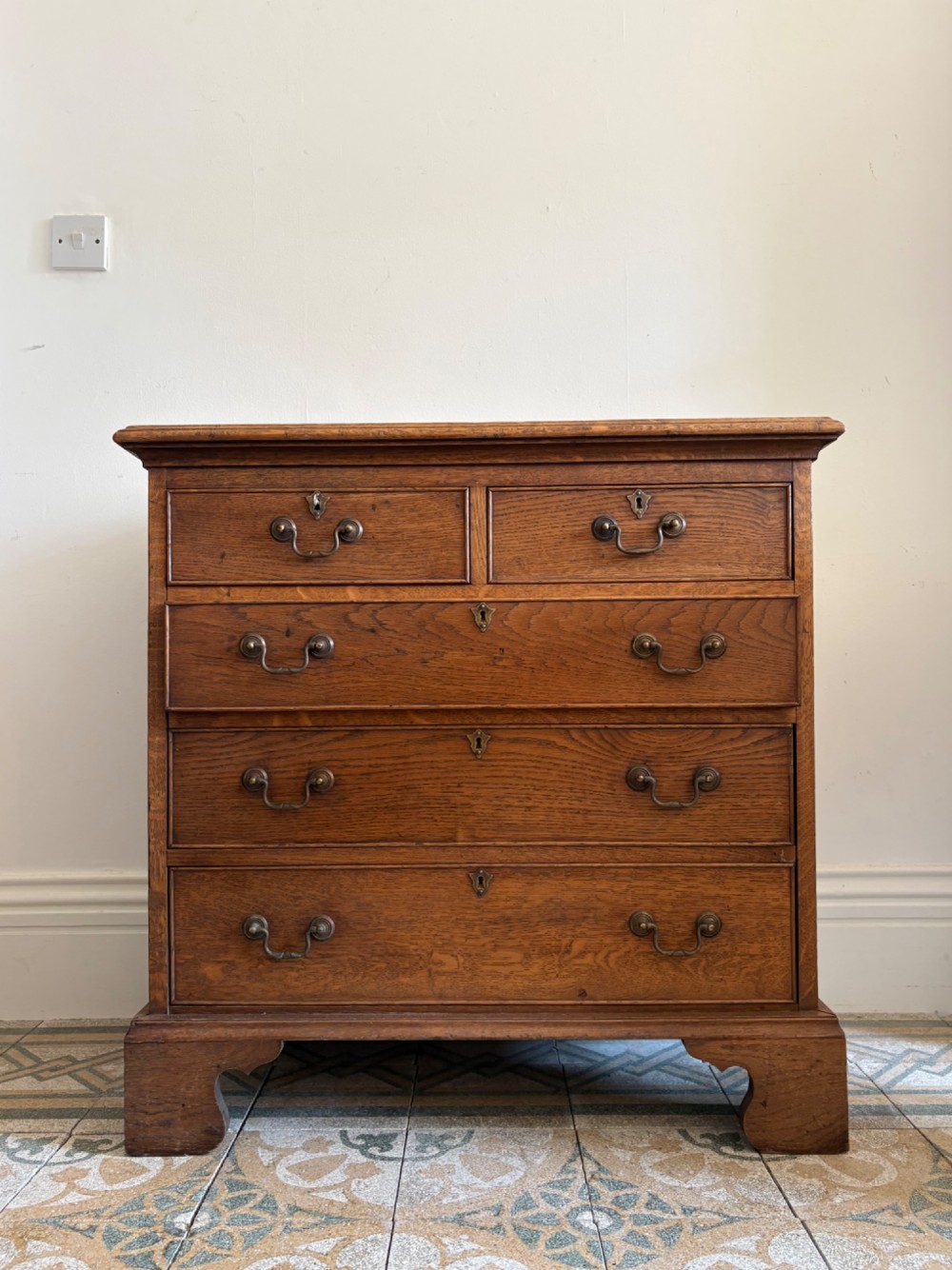 A George III style medium oak chest of drawers featuring two short drawers above three long drawers, resting on shaped bracket feet with brass handles.
