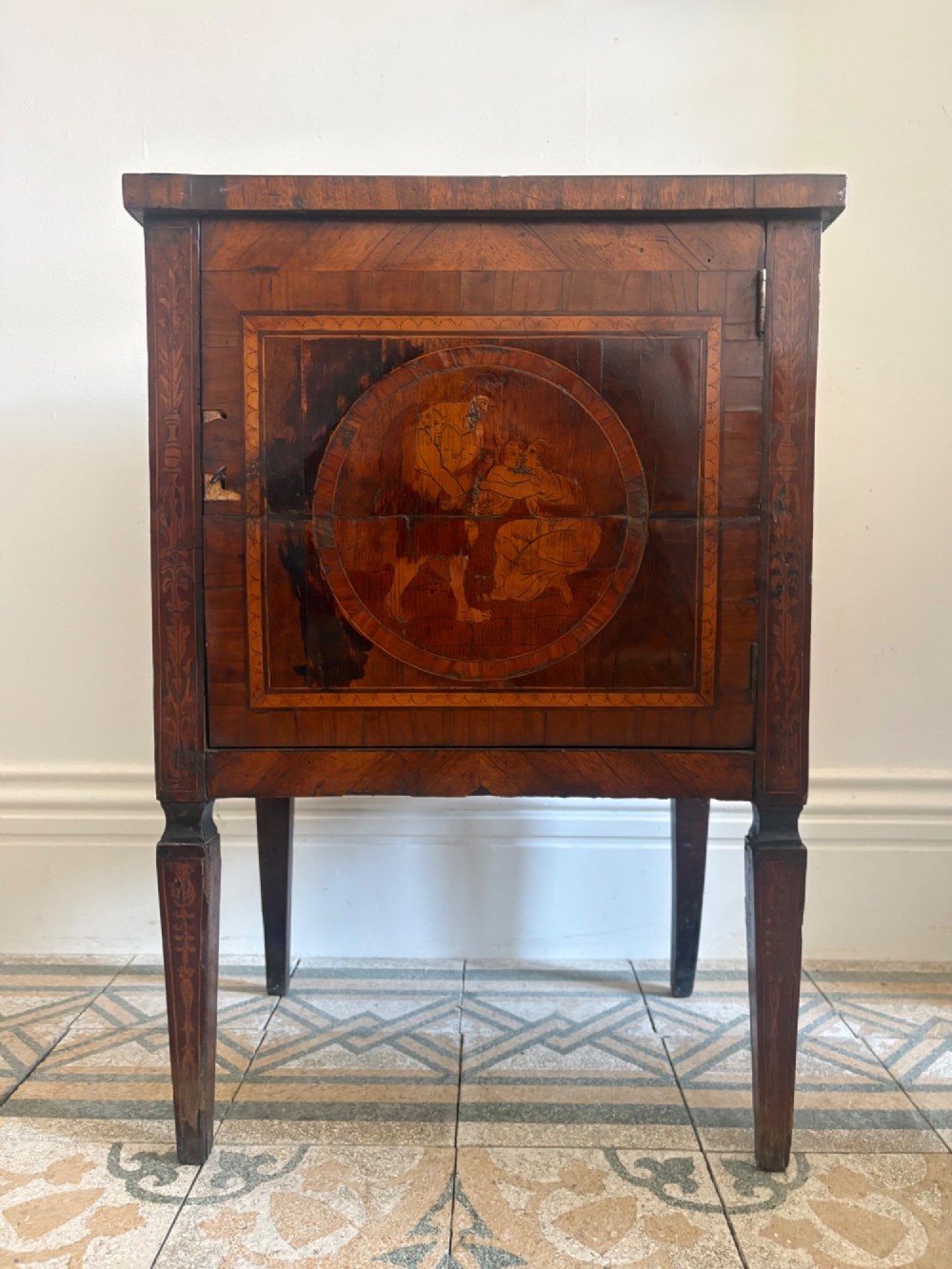 An 18th-century Italian Neoclassical walnut bedside cabinet featuring a central circular figurative marquetry panel on the front door, supported by square tapered legs.