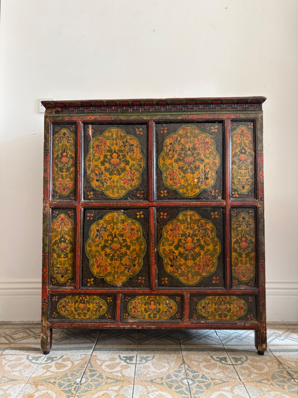 A 19th-century Tibetan wooden cabinet featuring four central hand-painted floral panels framed in red and gold mineral pigments.