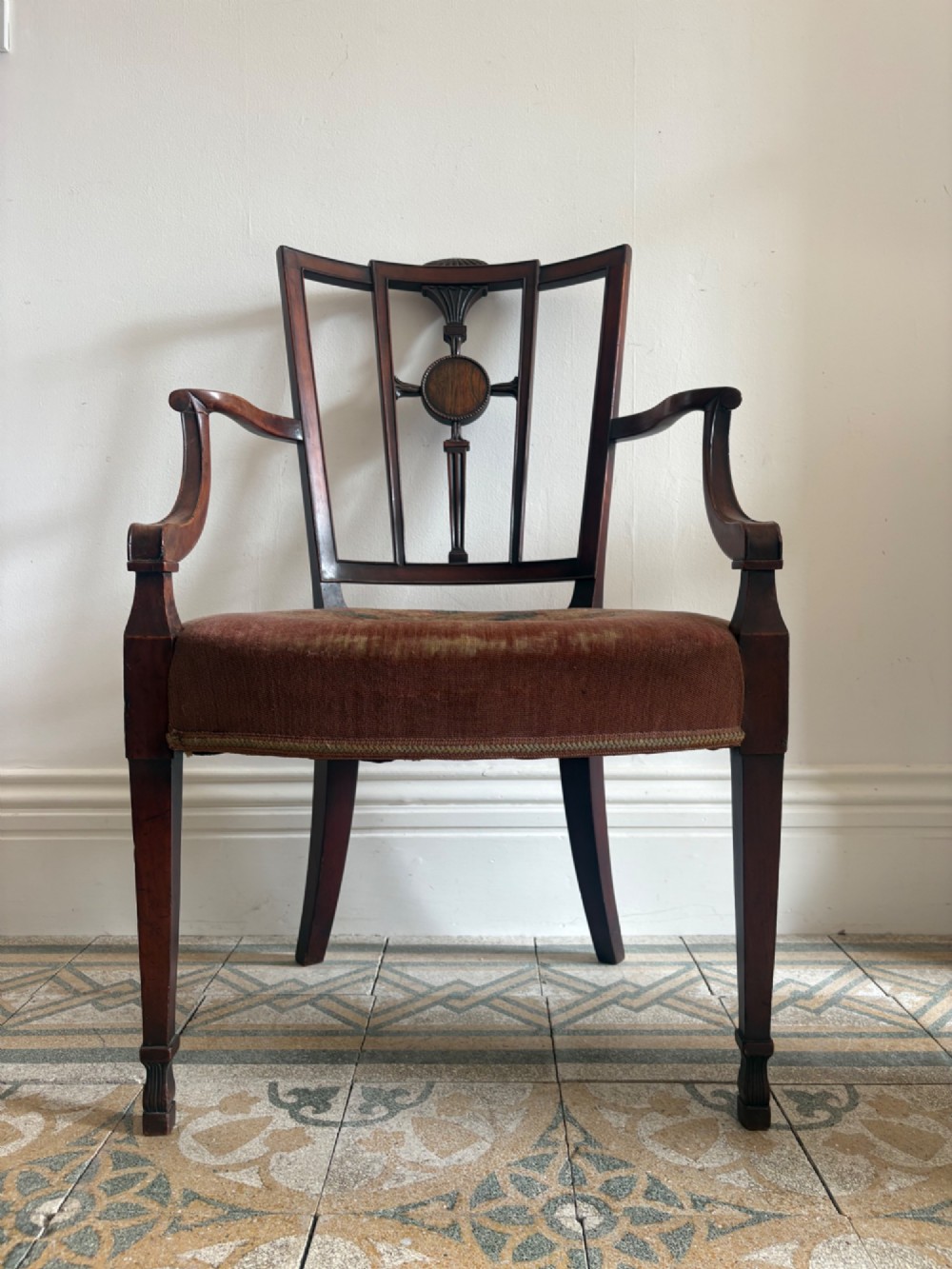 A late 18th-century Georgian mahogany open armchair featuring a carved back splat with a central rosewood roundel, swept armrests, and reddish-brown fabric upholstery.