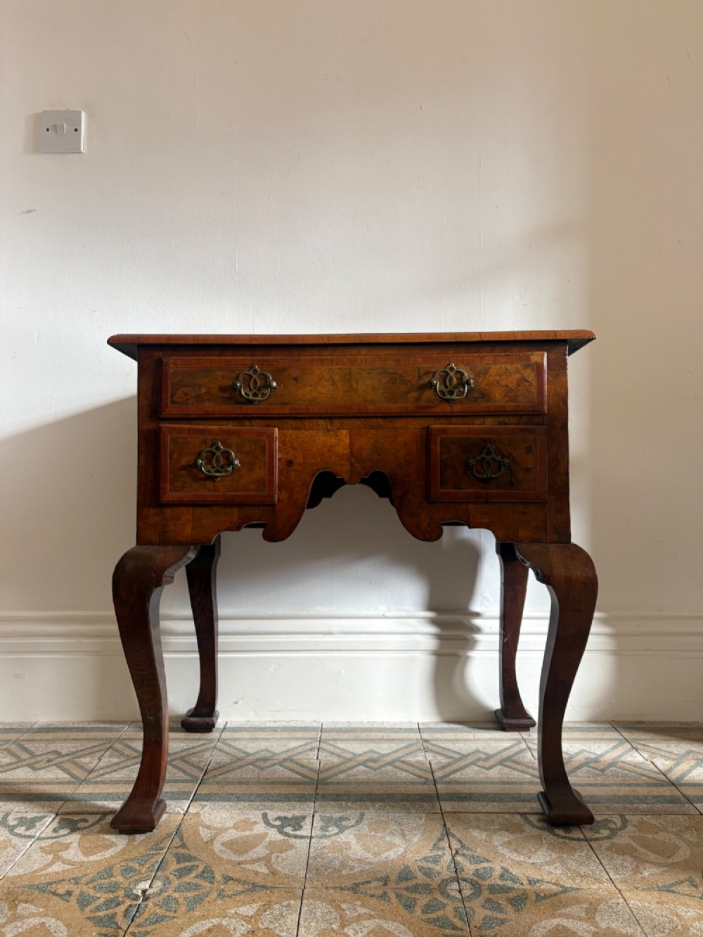 An early 18th-century figured walnut lowboy side table featuring three drawers, a shaped apron, brass handles, and cabriole legs.