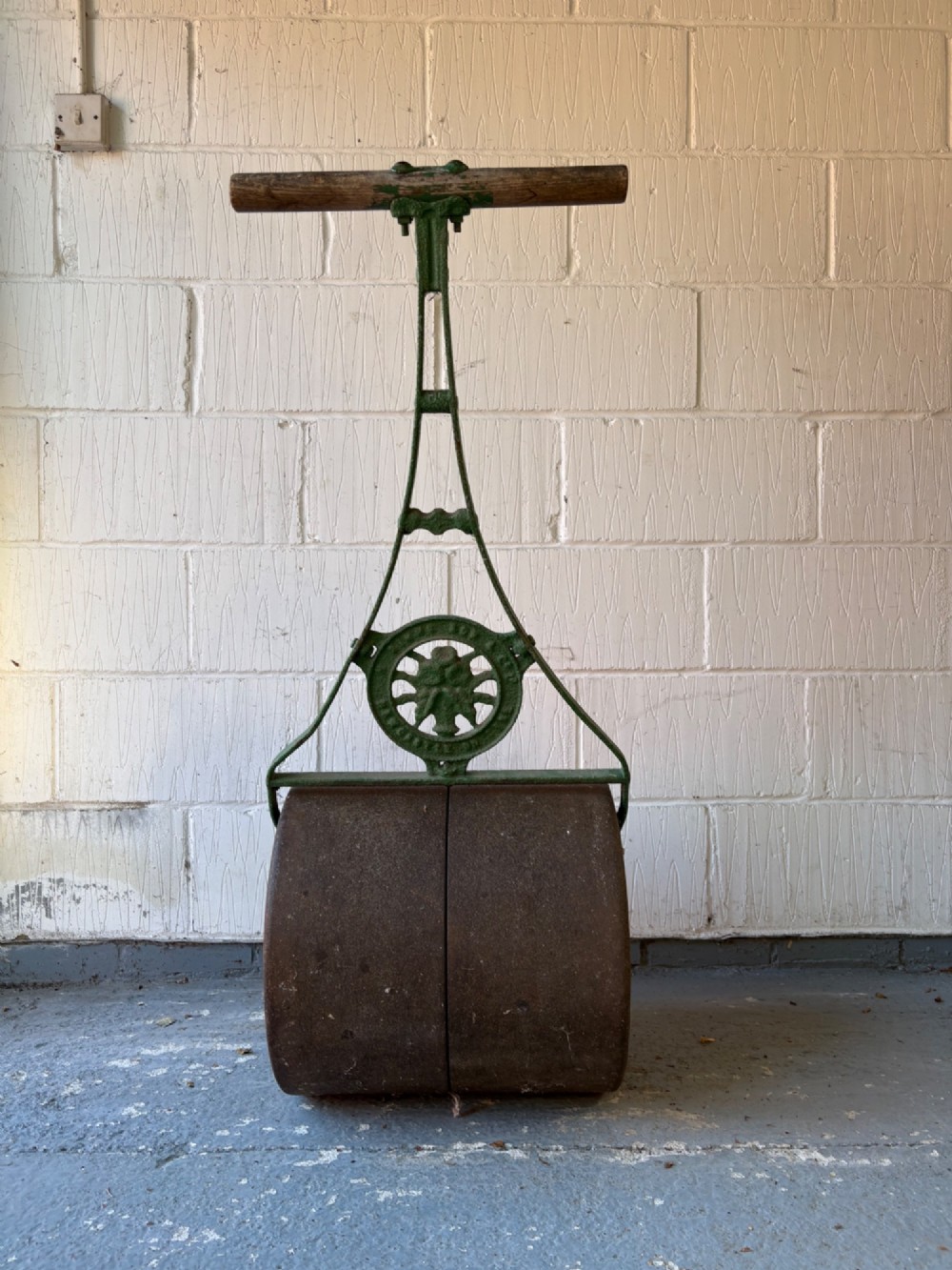 An antique cast iron garden roller featuring a green painted openwork frame with circular crest, a split drum cylinder, and a wooden handle bar. View III