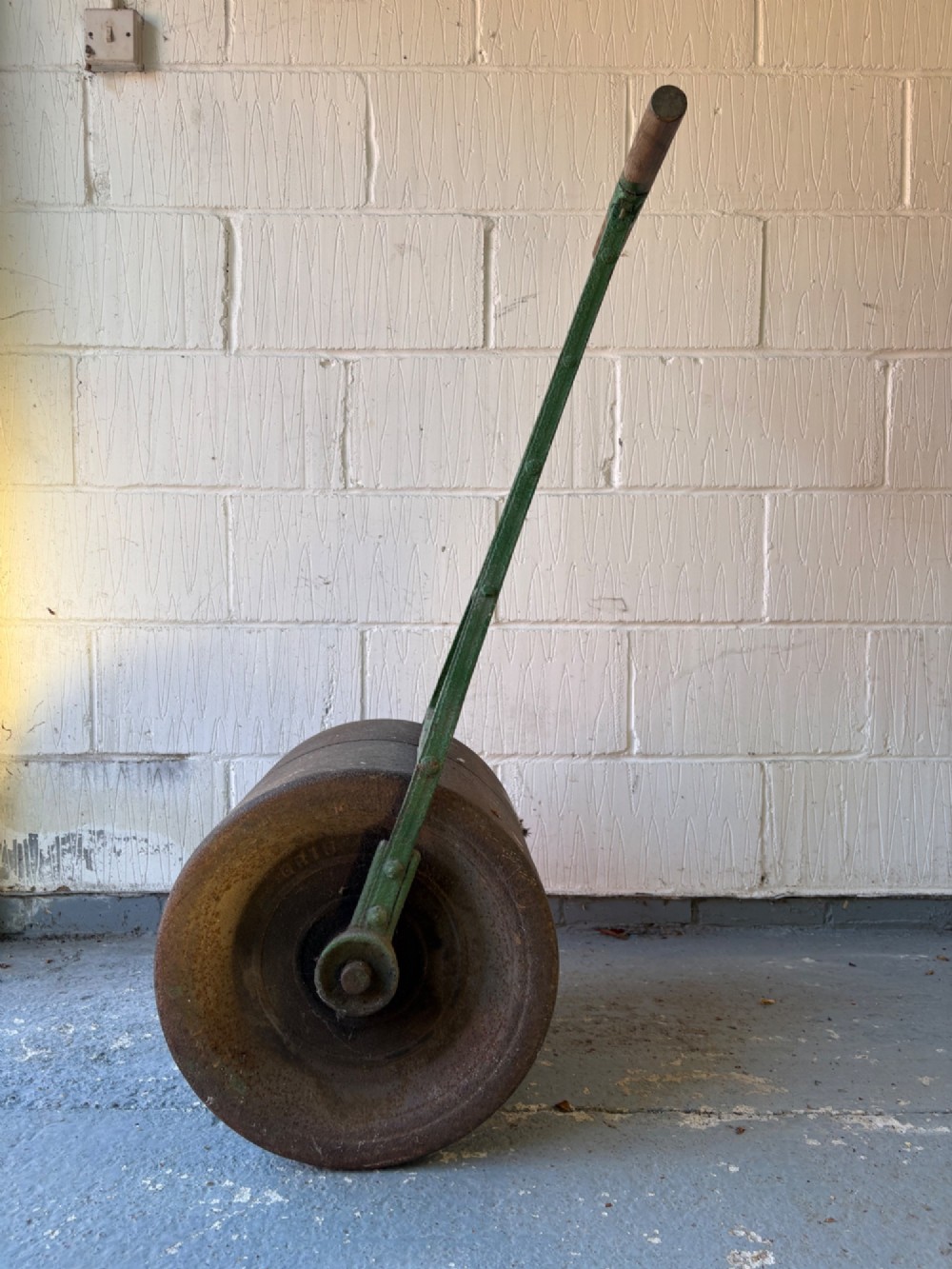 An antique cast iron garden roller featuring a green painted openwork frame with circular crest, a split drum cylinder, and a wooden handle bar. View IV