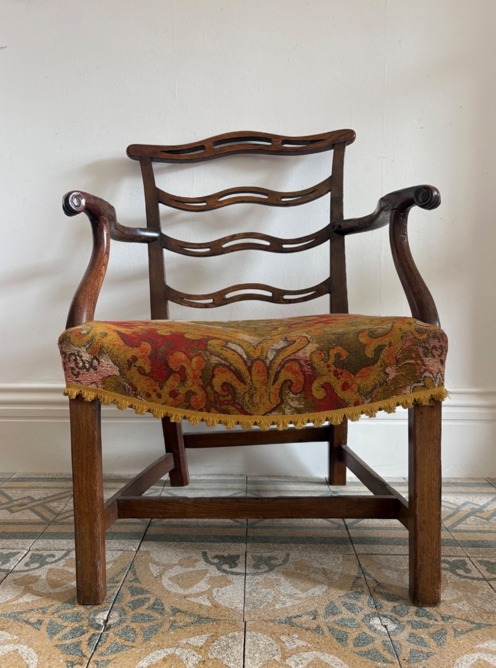 A George III elm ladderback open armchair featuring scrolled armrests and a patterned upholstered seat with fringed trim.